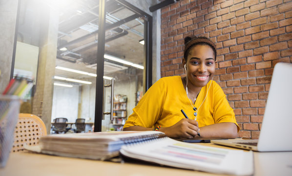 Portrait of beautiful smile black girl read books listen to earphones in library campus or modern office. Young creative casual business woman working on desk with computer