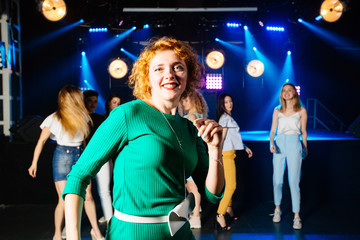 Pretty mature red haired curly female clubber dancing with her friends on background and looking at camera with smile during holidays relaxing celebrating at party in night club. Bright lighting.