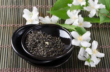 Closeup of dry green tea and jasmine flowers on the black saucer, green bamboo serving mat