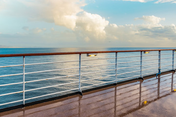 Wet cruise deck with railing and lights on a rainy day. White clouds and sea in the background.