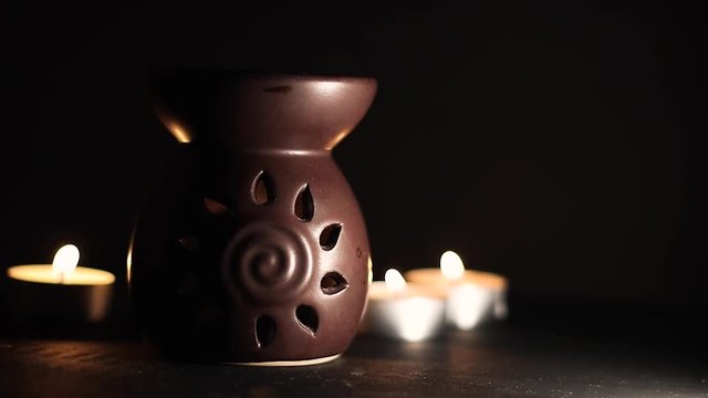 Traditional aroma diffuser with candle inside of it and candles in the back lighting up the dark scene, high contrast