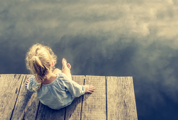 Lonely small girl dreaming on dock near small lake on warm summer day