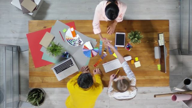 Directly above view of two interior designers standing at table and shaking hand of their female client standing in front of them and then continuing communication