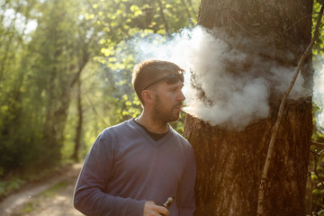 Vape man. An adult white bearded man in sunglasses smokes an electronic cigarette outside in the forest in sunny day. Bad habit that is harmful to health.