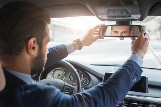 Everything should be perfect. Back view of young and handsome bearded businessman in formal wear adjusting the rearview mirror while driving his car.
