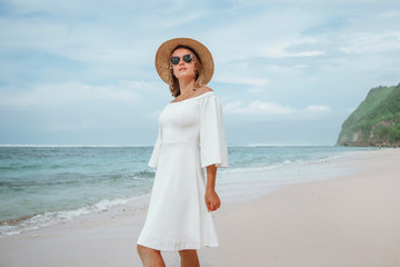 girl in a white dress walks on a white beach in a hat and sunglasses