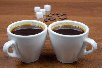 Two cups of espresso coffee on a wooden table with coffee beans and sugar in the background