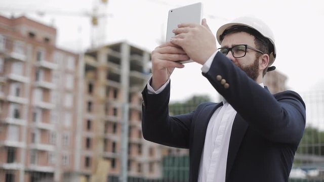 Young bearded architect at the construction site taking photo with tablet. He dressed in business suit.