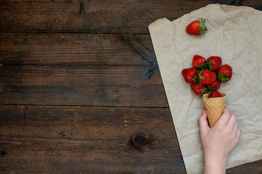 Children's hand takes strawberries in ice cream cone on natural wooden background. Strawberries in wafel cone and Baby girl hand. Summer vitamins banner