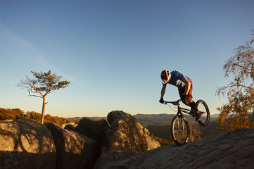 Fototapeta na wymiar Professional cyclist balancing on front wheel on trial bike. Sportsman rider making acrobatic trick on the edge of big boulder on the top of mountain. Concept of extreme sport active lifestyle