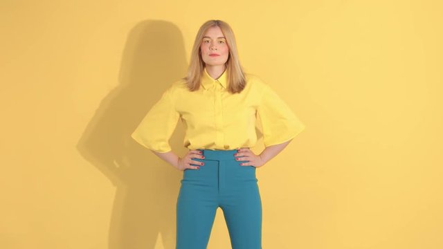 young woman in bright clothes on bright yellow background in studio watching straight to the camera with heand on waist. Hair blowing. Slow motion 60 fps