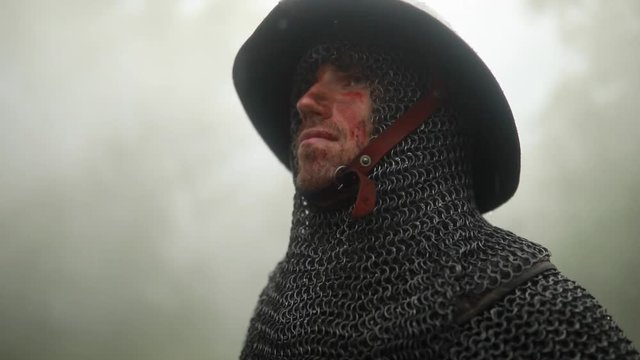 Portrait of medieval knight in armor with blood and wounds on his face on the background of the misty forest  in the rain.