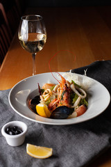 Seafood made dish with king prawns, mussels, squid, greens and citrus served with white wine and black olives on a white plate and wooden table