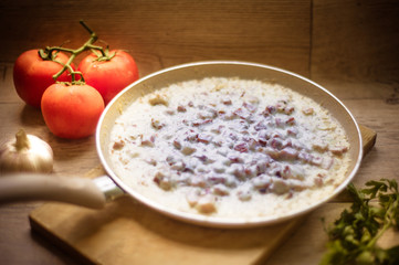White sauce in a frying pan served on a wooden plate on a kitchen counter, decorated with tomato. garlic and parsley
