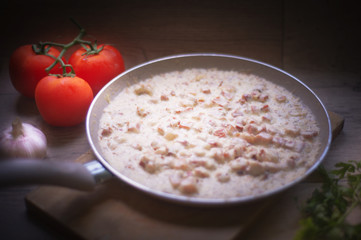 White sauce in a frying pan served on a wooden plate on a kitchen counter, decorated with tomato. garlic and parsley