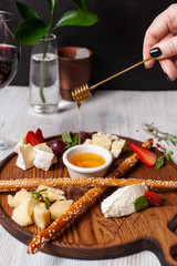 Female hand is pouring honey above assorted cheese: brie, gouda, honey, parmesan, camembert and strawberries, served on a wooden board with glass of red wine