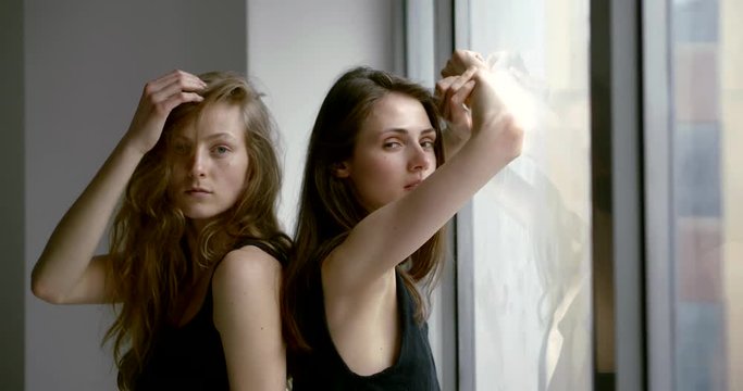 two pretty sisters are posing together near window, standing back to each other