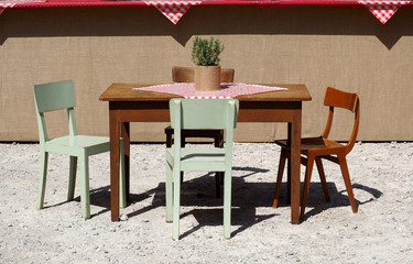 Rustic wooden table and colored chairs, with checkered tablecloth and rosemary plant, in a gravel front yard. Background for copy space, 