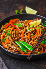 Vegan buckwheat soba noodles with vegetables in black plate on dark background.