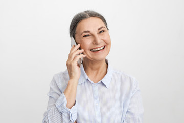 Studio shot of cheerful ecstatic senior elderly woman wearing elegant blue shirt laughing at good funny joke, showing her perfect straight teeth, enjoying nice phone conversation using mobile