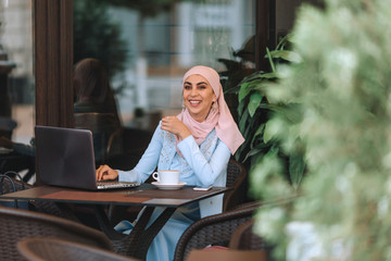 Muslim business woman working on a laptop in a cafe with a cup of coffee and chewing.