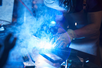 Close-up of busy welder in protective mask and gloves joining metal pieces with welding torch in...