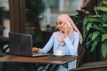 Muslim business woman working on a laptop in a cafe with a cup of coffee and chewing.