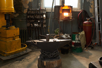 Workplace in smithy: steel anvil with hammer and tongs, heated furnace, gas tank and forging press in background