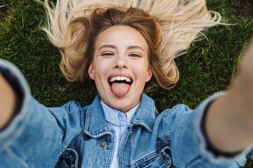 Photo of happy adorable woman taking selfie photo and showing her tongue while lying green grass in park © Drobot Dean
