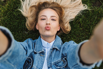 Photo of happy kind woman taking selfie photo and sending kiss while lying green grass in park