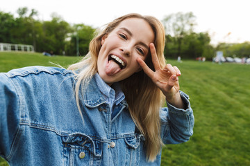 Photo of joyful smiling woman taking selfie photo and gesturing peace sing while walking in green park