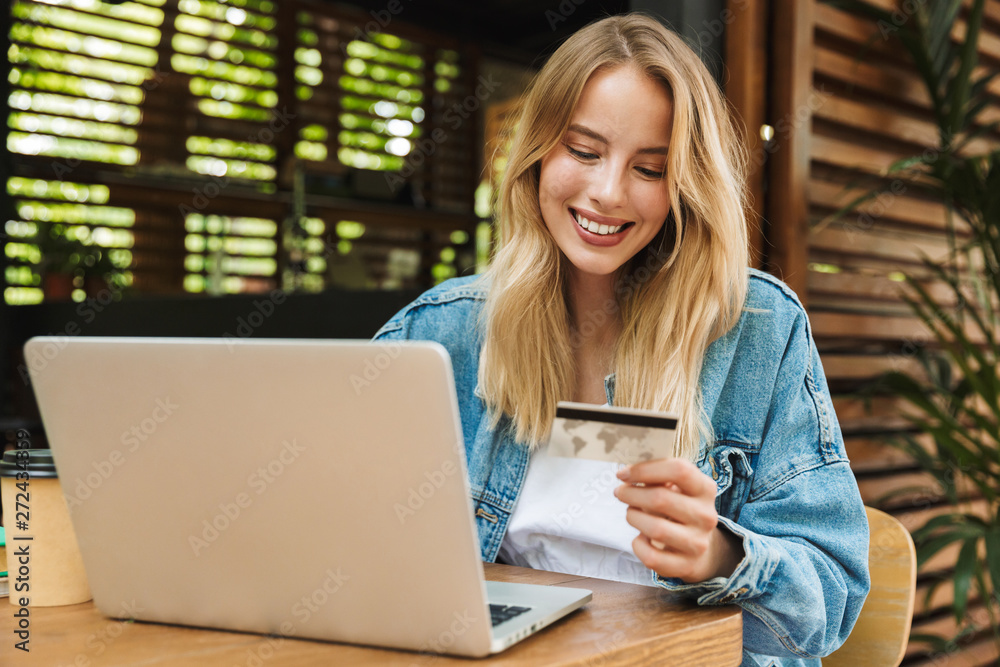 Sticker portrait of joyful young woman holding credit card while using laptop in cafe outdoors