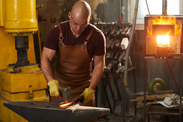 Focused young man in leather apron and protective gloves standing at anvil and beating hot iron...