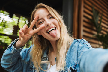 Portrait of pleased attractive woman taking selfie photo and showing peace sing in cafe outdoors