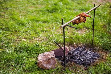Roast chicken with bamboo skewer On the fireplace from wood firewood and campfire charcoal in the blurred forest, the background is green grass.Retro style shallow focus effect.