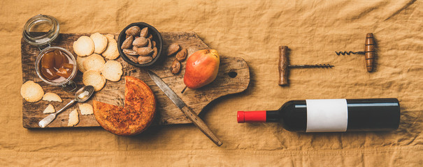 Wine and snack set. Flat-lay of wine bottle with blank label, vintage corkscrews and cheese and appetizers board over dusty yellow linen tablecloth, top view, wide composition. Party food concept