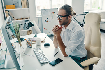 Concidering next step. Top view of thoughtful young man in shirt working using computer while sitting in the office