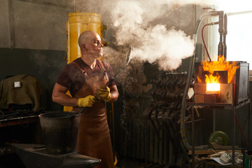 Serious bald young male metalworker in leather apron blowing on forged knife in smoke while...