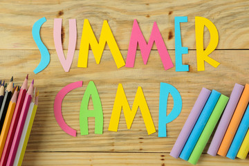 Text SUMMER CAMP of multicolored paper letters and color pencils on a natural wooden background. top view. flat lay