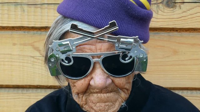 Gangster granny. Clouse up.  Grannie's greeting to the world. Grandmother is smiling. She is wearing a cool sun glasses in form of pistols and hat. Ecological background.