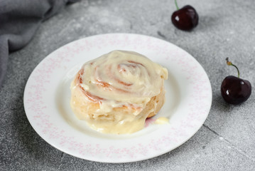 Cinnamon bun on a round plate on a gray background, decorated with a linen towel and cherry. Horizontal image.