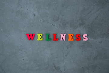 The multicolored wellness word is made of wooden letters on a grey plastered wall background.