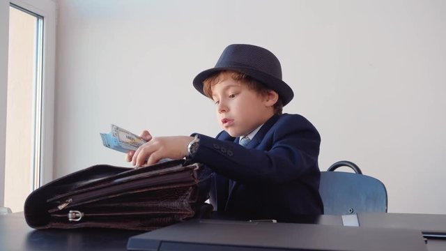 Little boy in suit and hat looks like entrepreneur is sitting in his office. He is hiding the cash money to briefcase and do paper work.