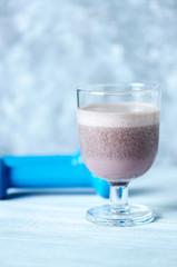 Glass of Chocolate Protein Shake with milk and black currant . Blue dumbbells in background. Sport nutrition. Wooden background. Copy space. 