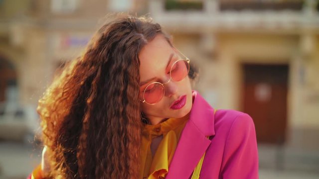 Outdoor close up portrait of fashionable woman with freckled skin, long curly hairs, wearing pink round sunglasses, colorful clothes, posing in street