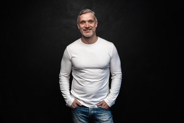 Middle-aged good looking man in white t-shirt posing in front of a black background with copy space.