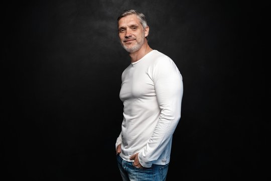 Middle-aged good looking man in white t-shirt posing in front of a black background with copy space.