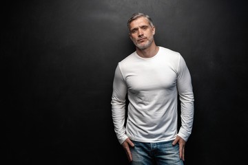 Middle-aged good looking man in white t-shirt posing in front of a black background with copy space.