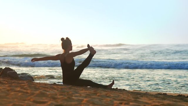 girl silhouette in black summer tracksuit shows pose stretching leg on sand beach against ocean under blue sky extreme slow motion. Concept yoga teacher guru group healthy lifestyle