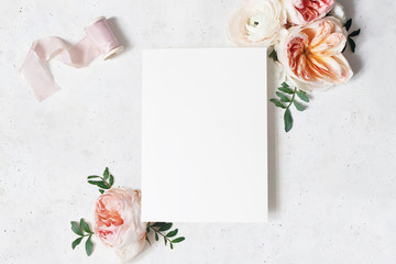 Feminine wedding, birthday stationery mock-up scene. Blank paper greeting card with bouquet of green leaves, blush pink English roses and ranunculus flowers. Concrete table background. Flat lay, top